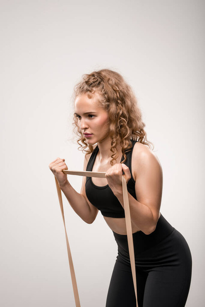 Fit sportswoman em traje preto dobrando ligeiramente para a frente durante o exercício para o braço e usando banda flexível durante o treinamento
 - Foto, Imagem