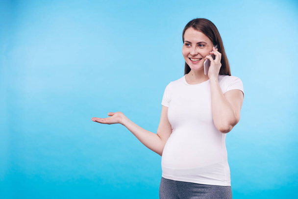 Smiling pregnant woman with smartphone by ear talking to her friend or husband while explaining or discussing something in isolation - Foto, imagen