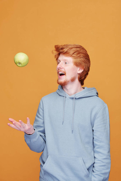 Happy young casual man throwing green apple in front of himself while having fun in front of camera over yellow background in isolation - Fotó, kép
