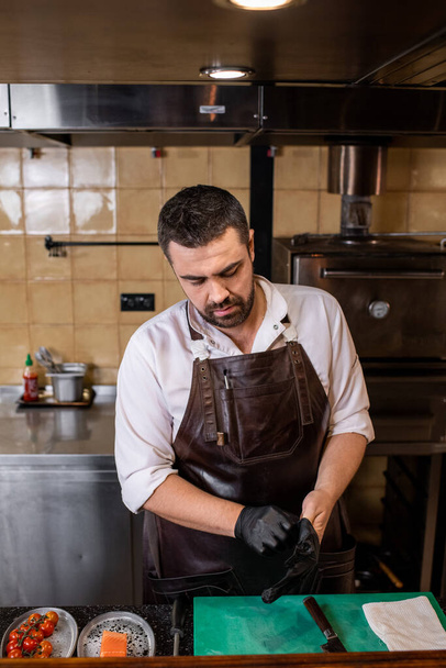 Chef barbudo ocupado en delantal de cuero de pie en el mostrador y poner guantes en las manos antes de cocinar - Foto, Imagen