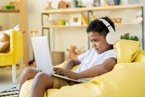Concentrated adorable schoolkid in headphones looking at laptop display while playing game or taking online educational course - Fotografie, Obrázek