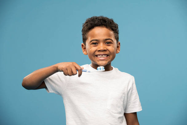 Glücklicher süßer afrikanischer oder gemischter Rasse kleiner Junge mit Zahnbürste, der seine Zähne putzt, während er vor der Kamera auf blauem Hintergrund steht - Foto, Bild