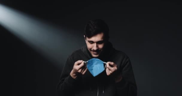 Portrait shot of Caucasian young happy male patient taking off blue medical mask and smiling at camera with joyful face. Black wall background. Guy during coronavirus outbreak. - Filmati, video