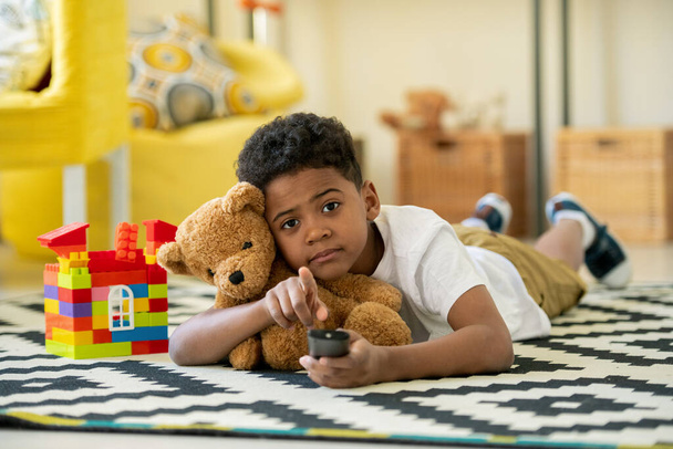 Cute African preschooler with remote control and brown teddybear pointing at you while lying on the floor of living-room - Foto, Bild