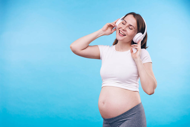 Young pregnant woman with headphones singing one of her favorite songs with cheering music while standing in front of camera - Photo, Image