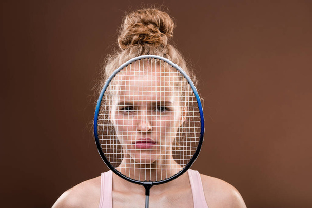 Face of young serious sportswoman with blond hair looking at you from behind tennis racket while standing in front of camera - Fotoğraf, Görsel