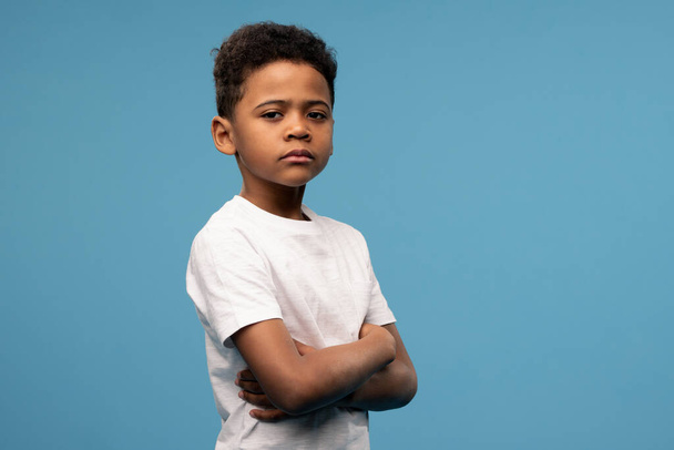 Serious or offended cute little boyin white t-shirt crossing his arms on chest while standing in front of camera over blue background - Photo, Image