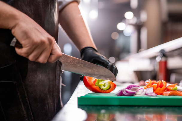 Gros plan d'un chef méconnaissable dans un tablier coupant du poivron avec un couteau de cuisine tout en faisant une salade d'été rafraîchissante - Photo, image