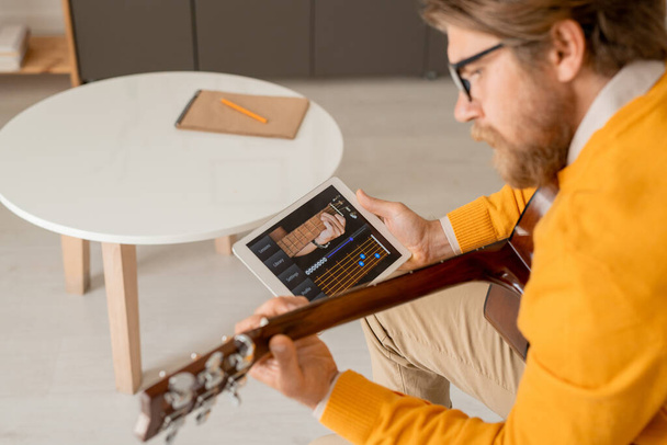 Joven hombre serio en puente amarillo inclinándose sobre su guitarra mientras usa touchpad para buscar reglas de afinación de instrumentos musicales - Foto, Imagen
