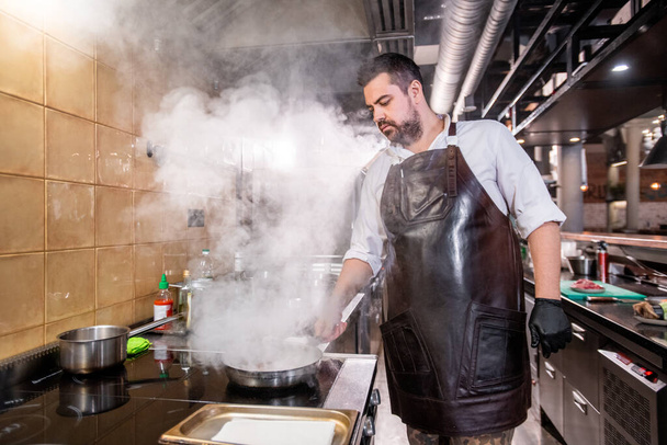 Chef barbu sérieux dans le tablier debout à la cuisinière et les ingrédients de cuisson sur la poêle, vapeur au-dessus de la poêle - Photo, image
