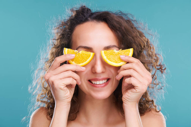 Jeune femme en bonne santé avec le sourire de dent et les cheveux foncés ondulés tenant deux moitiés de tranche d'orange juteuse par ses yeux pendant la procédure de beauté - Photo, image
