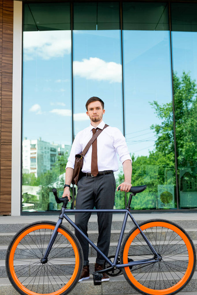 Joven hombre de negocios elegante en pantalones, camisa y corbata sosteniendo la bicicleta mientras está de pie en la escalera contra el exterior del edificio moderno - Foto, imagen