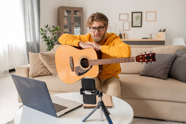 Young music teacher talking to his audience during online lesson of playing guitar while sitting on couch in front of smartphone camera - Foto, Imagem
