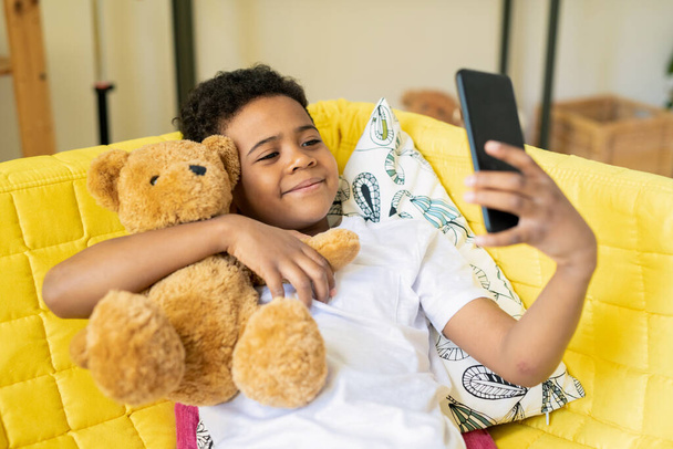 Щасливий хлопчик африканської етнічної приналежності зі смартфоном, який обіймає свого плюшевого ведмедика, сидячи на жовтому дивані і роблячи самовідтворення. - Фото, зображення