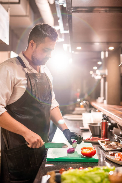 Serieuze bebaarde chef-kok werkt in de commerciële keuken en snijdt rode ui, lens flare - Foto, afbeelding