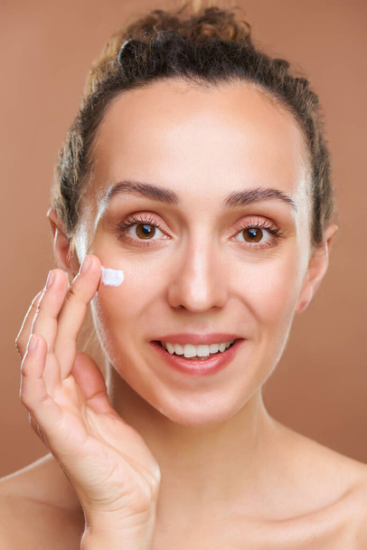 Šťastná a zdravá mladá žena nanášení hydratační krém na její obličej, zatímco stojí před kamerou na hnědém pozadí - Fotografie, Obrázek