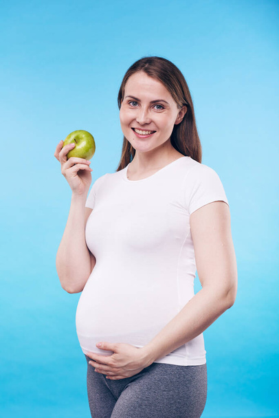 Happy pregnant female with granny smith apple touching bottom of her belly while standing in front of camera in isolation - Фото, зображення