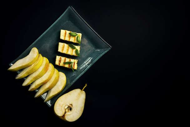 Grilled slices of halloumi cheese on a plate with slices of pear and Basil leaf, on a dark background. Traditional cheese of Greece and Turkey. Copy space. flat lay. - Fotoğraf, Görsel