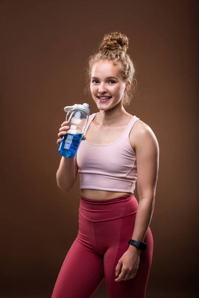 Молодая веселая спортсменка с зубастой улыбкой держит пластиковую бутылку с освежающим напитком во время перерыва после тренировки в тренажерном зале - Фото, изображение