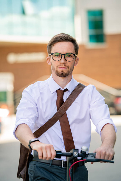 Young serious businessman in eyeglasses and formalwear going home by bicycle while moving forwards in front of camera outdoors - Photo, Image