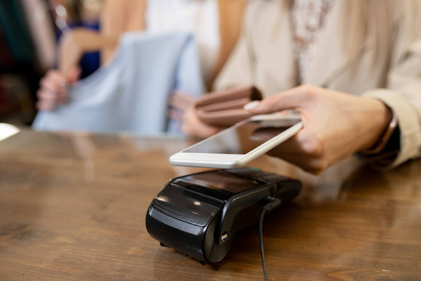 Main de jeune femme shopaholic tenant smartphone sur la machine de paiement sur le comptoir tout en achetant de nouveaux vêtements en boutique - Photo, image