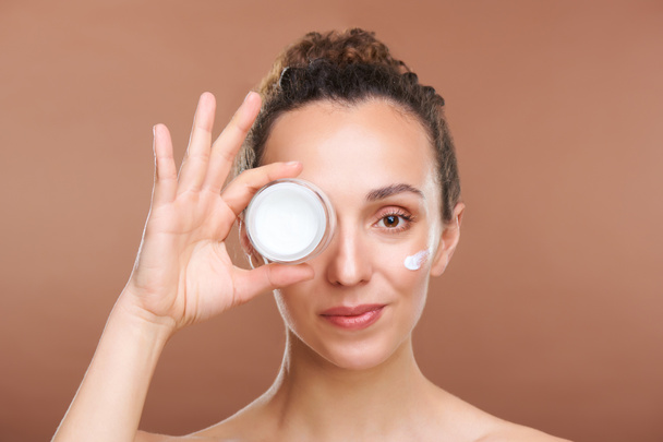 Healthy young beautiful woman holding small jar of rejuvenating facial cream by eye while standing in front of camera in isolation - Фото, зображення