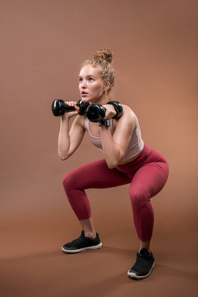 Молода сильна спортсменка в спортивному костюмі сидить на присіданнях і піднімає гантелі, докладаючи зусиль під час важких вправ
 - Фото, зображення