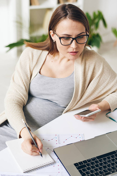 Ziemlich schwangere Analytikerin oder Maklerin mit mobilen Geräten, während sie Online-Informationen mit denen in Finanzgrafiken nach Arbeitsplatz vergleicht - Foto, Bild