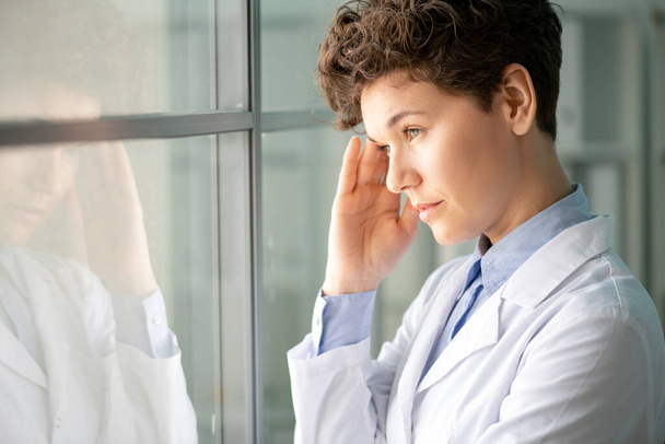 Trabalhadora de laboratório cansada com cabelo curto em pé na janela da sala de experiências e no templo de massagens enquanto sente stress
 - Foto, Imagem