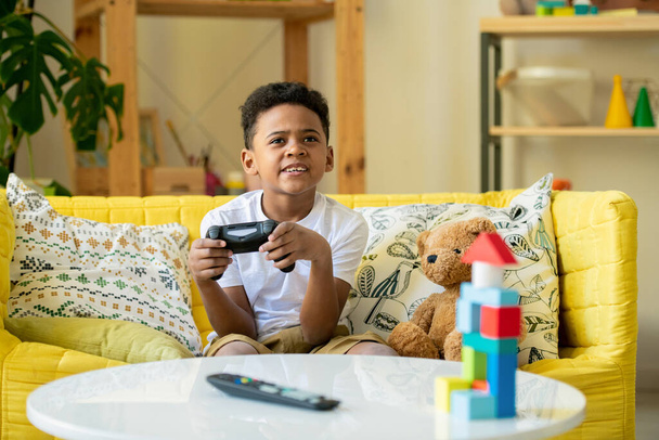 Little African or mixed-race boy with joystick looking at tv screen while sitting on yellow couch by table and playing video game - Photo, Image