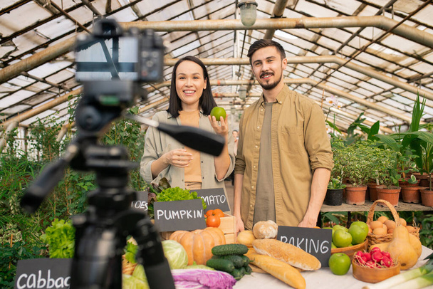 Positives junges multiethnisches Paar zeigt Apfel vor der Kamera, während es Video-Review im Gewächshaus dreht - Foto, Bild