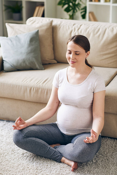 Молодая положительная беременная женщина в активной одежде сидит в позе лотоса за диваном на полу и медитирует, оставаясь дома - Фото, изображение