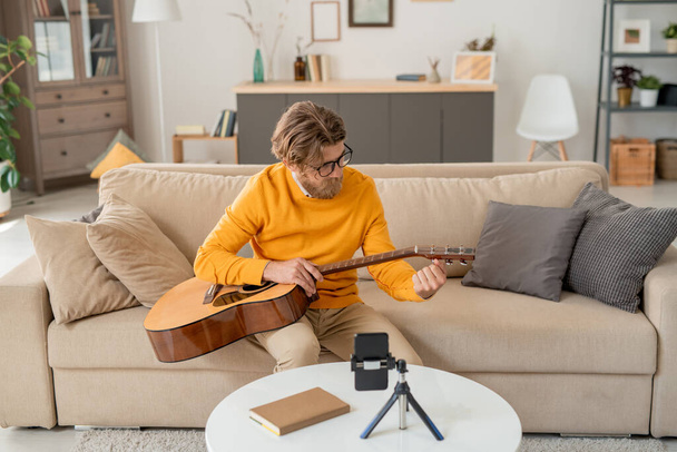 Joven contemporáneo en jeans y jersey amarillo sentado en el sofá y tirando de cuerdas de guitarra delante de la cámara del teléfono inteligente en casa - Foto, imagen