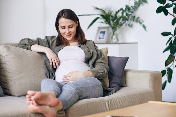 Junge barfüßige Frau in Freizeitbekleidung blickt auf ihren schwangeren Bauch, während sie es sich auf der Couch zwischen Kissen im Wohnzimmer gemütlich macht - Foto, Bild