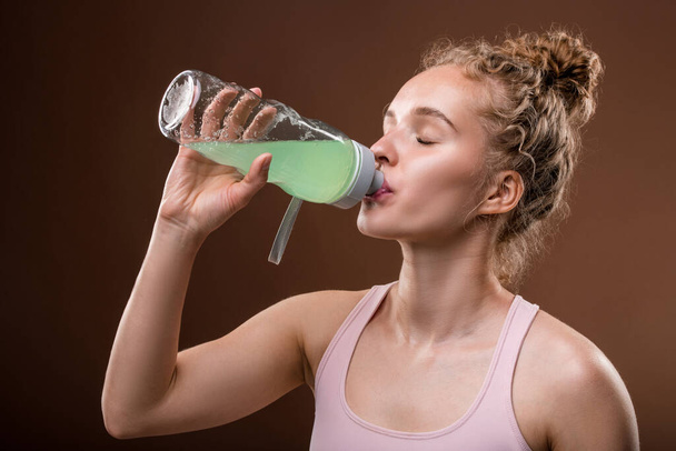 Mujer joven sedienta activa con cabello rubio rizado tomando una bebida refrescante de botella de plástico después de entrenar en el gimnasio o centro de lesiones - Foto, Imagen