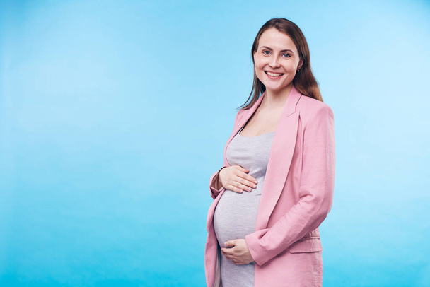 Happy young elegant future mother in grey casual dress and pink trenchcoat keeping hands of her belly while standing in isolation - Фото, изображение