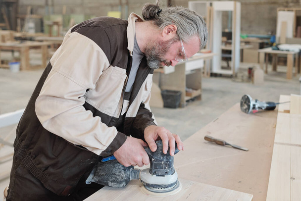 Lavoratore maturo di mobili che producono fabbrica piegandosi su tavola di legno durante l'utilizzo della smerigliatrice per levigare la superficie del pezzo - Foto, immagini