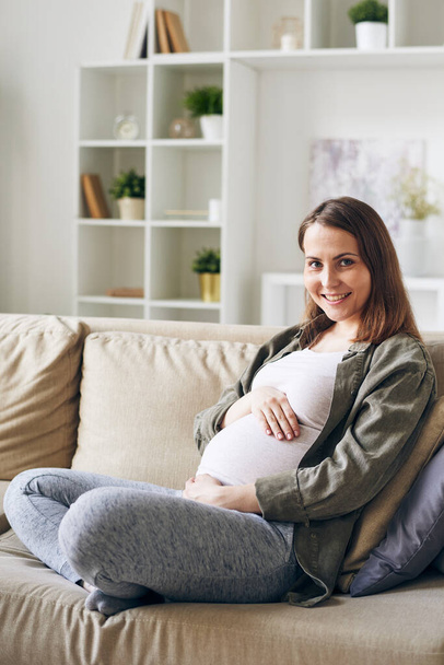 Boldog fiatal terhes nő alkalmi viseletben ül párnák között a kanapén a kamera előtt otthoni környezetben - Fotó, kép