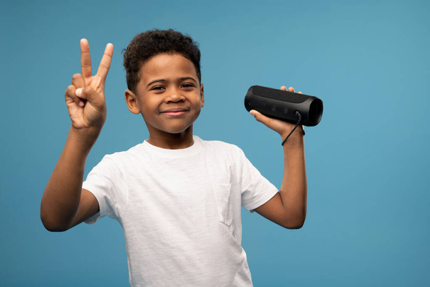 Веселый милый маленький мальчик африканской национальности, показывающий мирный жест во время веселья и прослушивания музыки в беспроводной интеллектуальной колонке
 - Фото, изображение