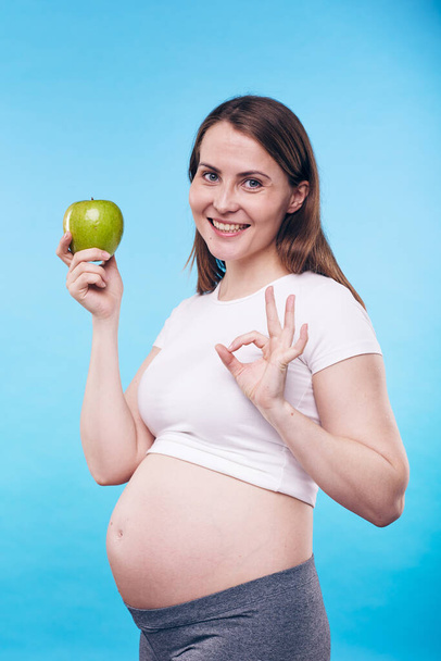 Happy and healthy young pregnant woman with green apple showing you okay gesture meaning that proper nutrition is important - Fotografie, Obrázek
