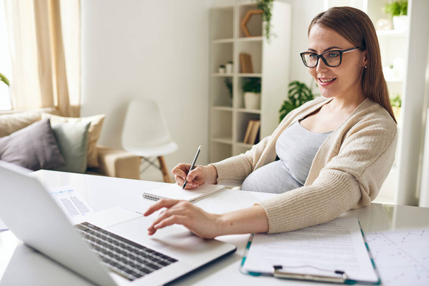 Счастливая молодая беременная деловая женщина, глядя на дисплей ноутбука и делая заметки в блокноте при анализе финансовой информации - Фото, изображение