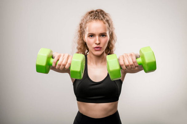 Young active sportswoman in black tracksuit standing in front of camera and looking at you while doing exercise with green dumbbells - Fotoğraf, Görsel