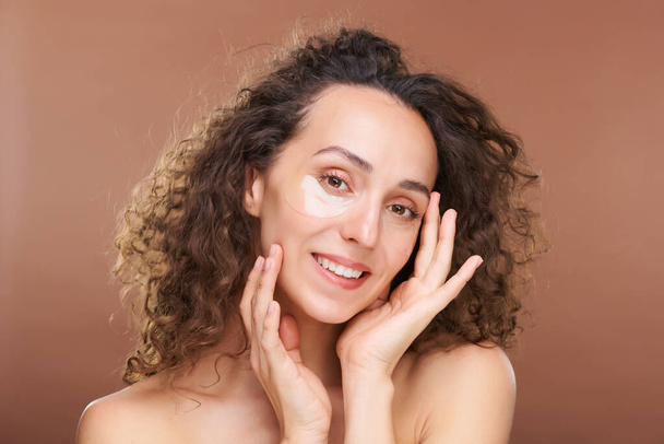 Jonge lachende vrouw met krullend donker haar en hydraterende vlekken onder de ogen aanraken van haar gezicht tijdens schoonheid procedure in isolatie - Foto, afbeelding