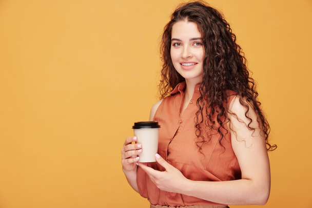 Felice giovane donna sorridente con lunghi capelli scuri ondulati in possesso di un bicchiere di caffè mentre in piedi davanti alla fotocamera con copyspace a sinistra - Foto, immagini