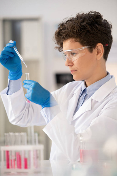 Microbiólogo joven concentrado en bata de laboratorio usando pipeta mientras deja caer el reactivo en el tubo de ensayo - Foto, imagen