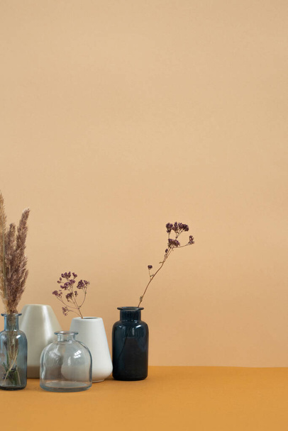 Группа керамических и стеклянных ваз с сушеными полевыми цветами и шипами, стоящих на столе напротив стены домашней комнаты или студии дизайна - Фото, изображение