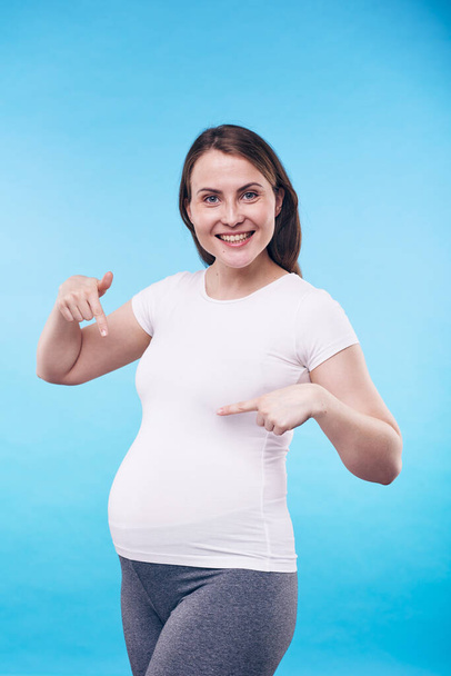 Весела вагітна жінка в активному одязі вказує на живіт і висловлює радість від очікування дитини на синьому фоні
 - Фото, зображення