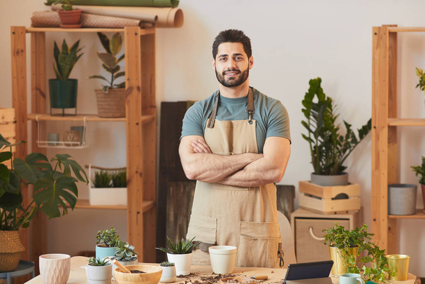 Waist up portrait of handsome bearded man wearing apron and looking at camera while standing by table with houseplants and gardening tools, copy space - Photo, Image