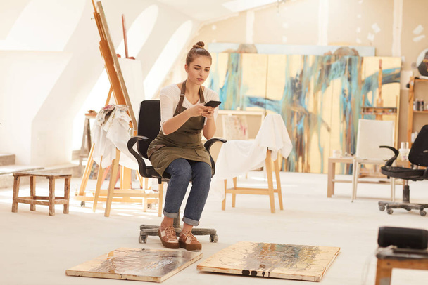Retrato de larga duración de una joven artista que toma fotos de pinturas mientras está sentada en una silla en un amplio estudio de arte iluminado por la luz del sol, espacio para copiar - Foto, imagen