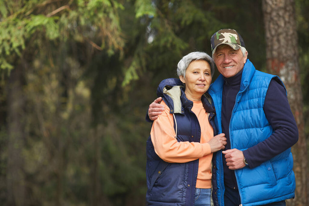 Талия портрет активной пожилой пары, обнимающей и смотрящей в камеру во время прогулки в лесу, копирующей пространство - Фото, изображение